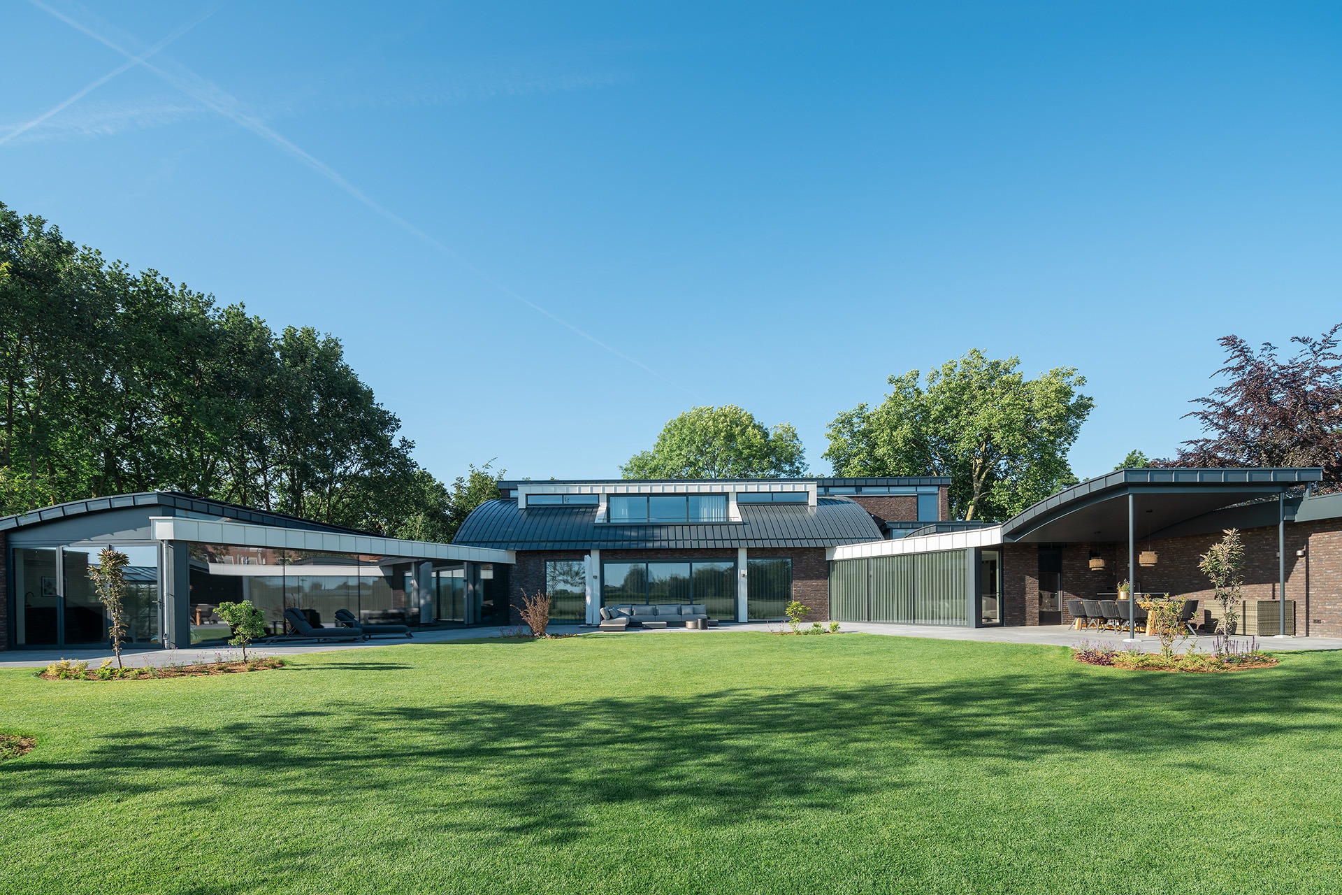 Achteraanzicht van Villa MB vanuit de achtertuin gezien. De achtertuin is een groot grasveld en aan het huis zitten twee terrassen waar zitplaatsen zijn gerealiseerd