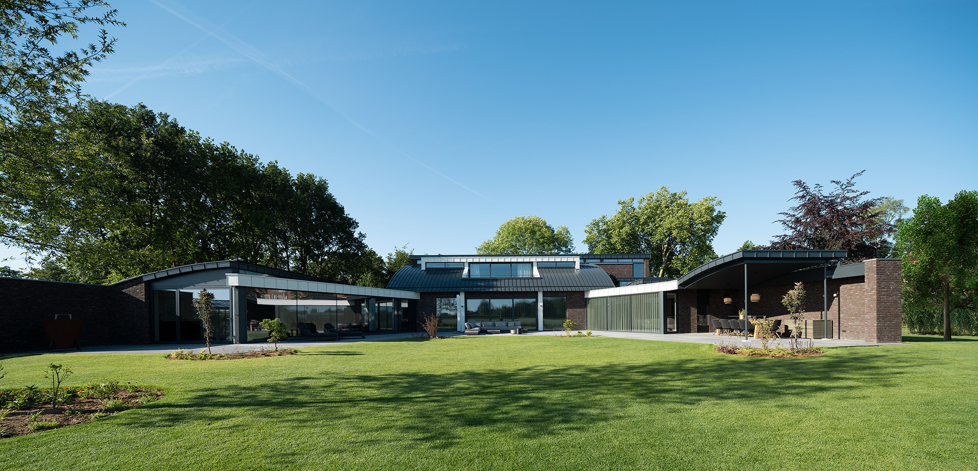 Achteraanzicht van Villa MB vanuit de achtertuin gezien. De achtertuin is een groot grasveld en aan het huis zitten twee terrassen waar zitplaatsen zijn gerealiseerd