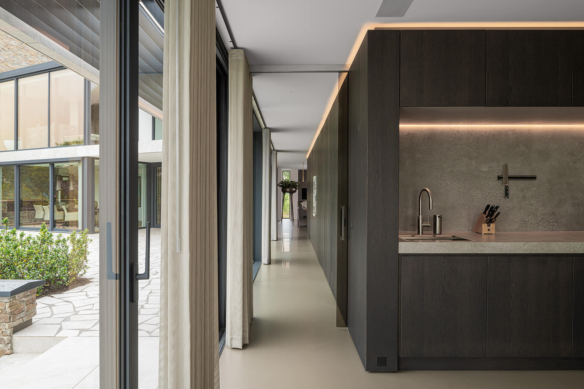 Detailfoto van de keuken en gang in Villa VS. Een lange gang met aan de linkerzijde gordijnen en aan de rechterzijde een grijs wandpaneel