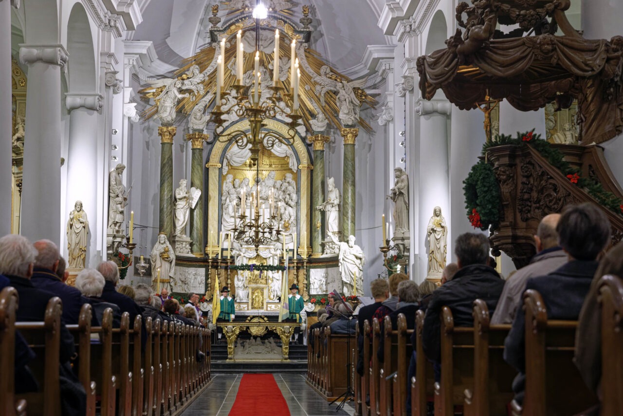 Binnenzijde van een volle Bartholomeuskerk. Op de voorzijde mensen in kerkbanken. Voor het altaar staan twee leden van het Gilde