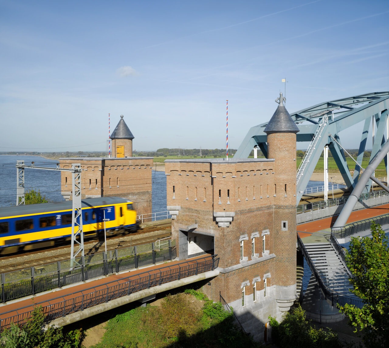 Een trein rijdt over de spoorbrug bij Zaltbommel, met op de voorgrond het gerestaureerde historische Landhoofd van baksteentorens.