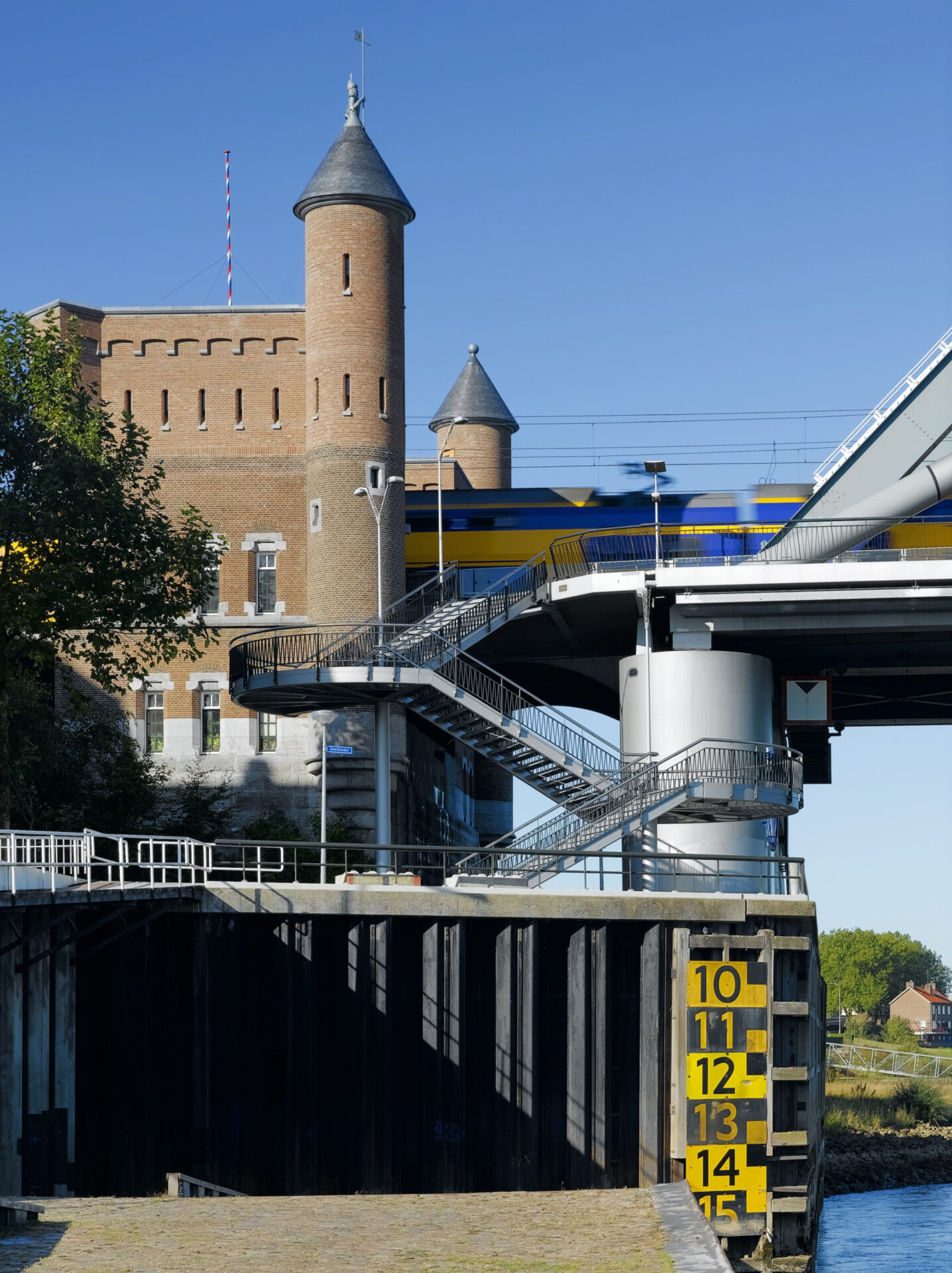 Het bakstenen landhoofd van de spoorbrug, met op de voorgrond een stalen trappentoren en een waterstandsmeter, terwijl een trein in over de brug passeert