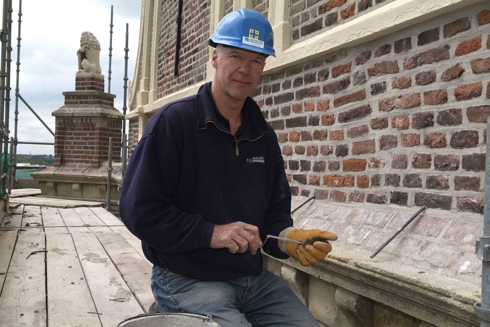 Portretfoto van metselaar van Van Dinther voor een klassiek gebouw. De metselaar draagt een helm en een handschoen
